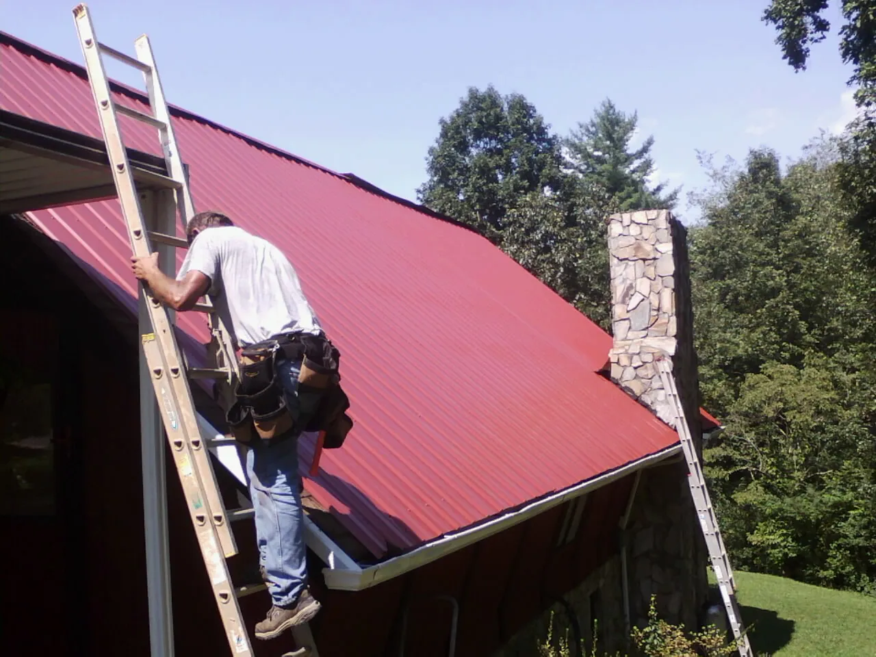 Roofer on a ladder beside a red metal roof and stone chimney — Soffit & Fascia Repair by Roofing Brothers in Pegram